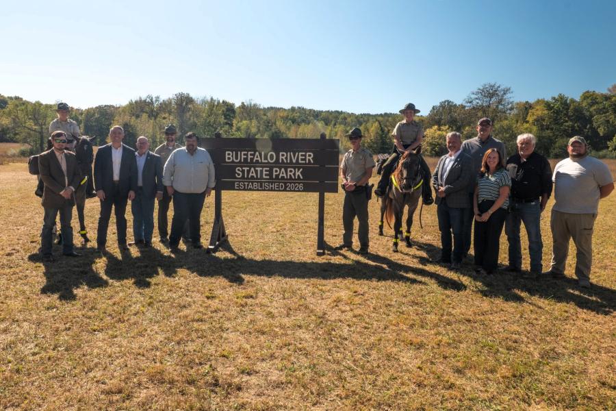 A group of people, including uniformed park rangers, pose beside a large wooden sign that reads "Buffalo River State Park – Established 2026." Two rangers sit on horses on either side of the sign. The group is in an open grassy field with trees and a clear blue sky in the background.